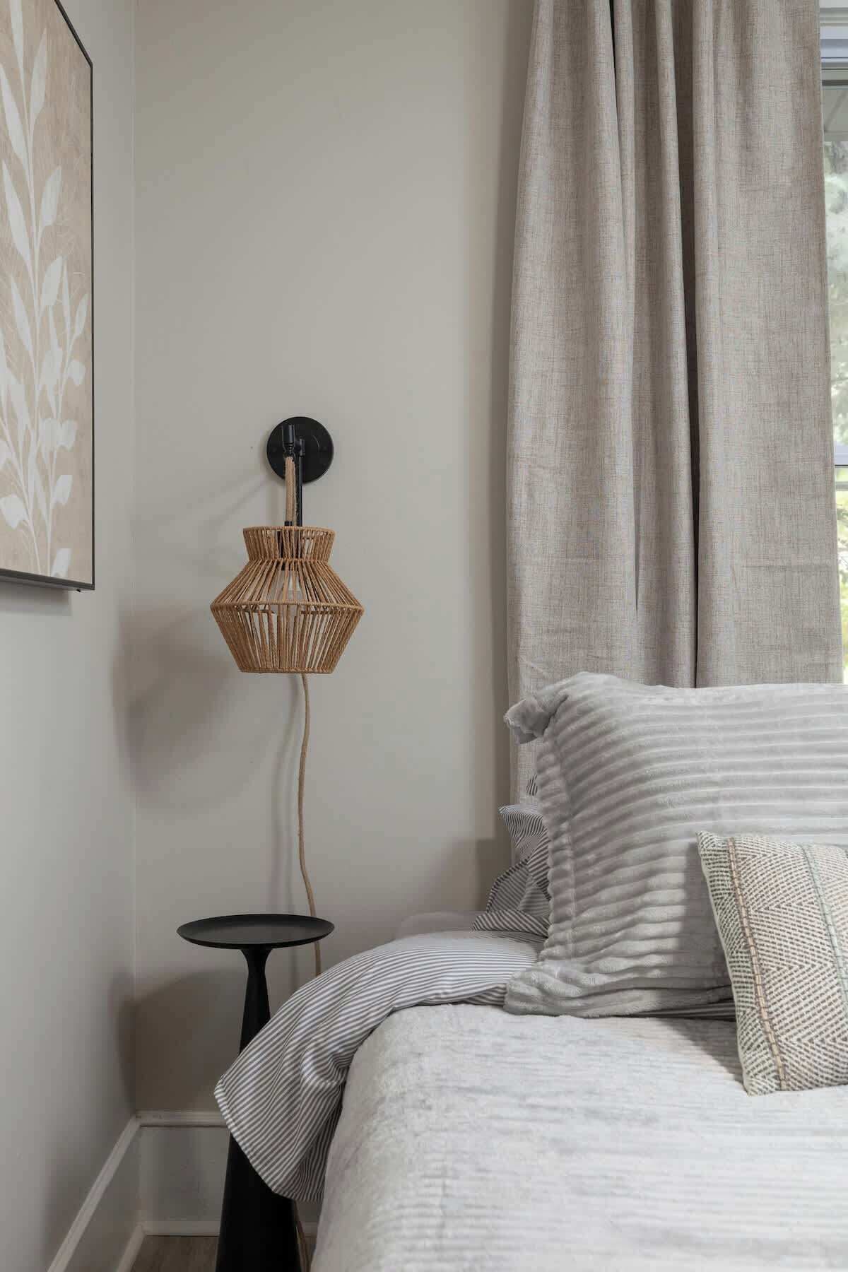 Une chambre à coucher avec un lit, une table de chevet, une lampe fixée au mur et une fenêtre avec des rideaux.