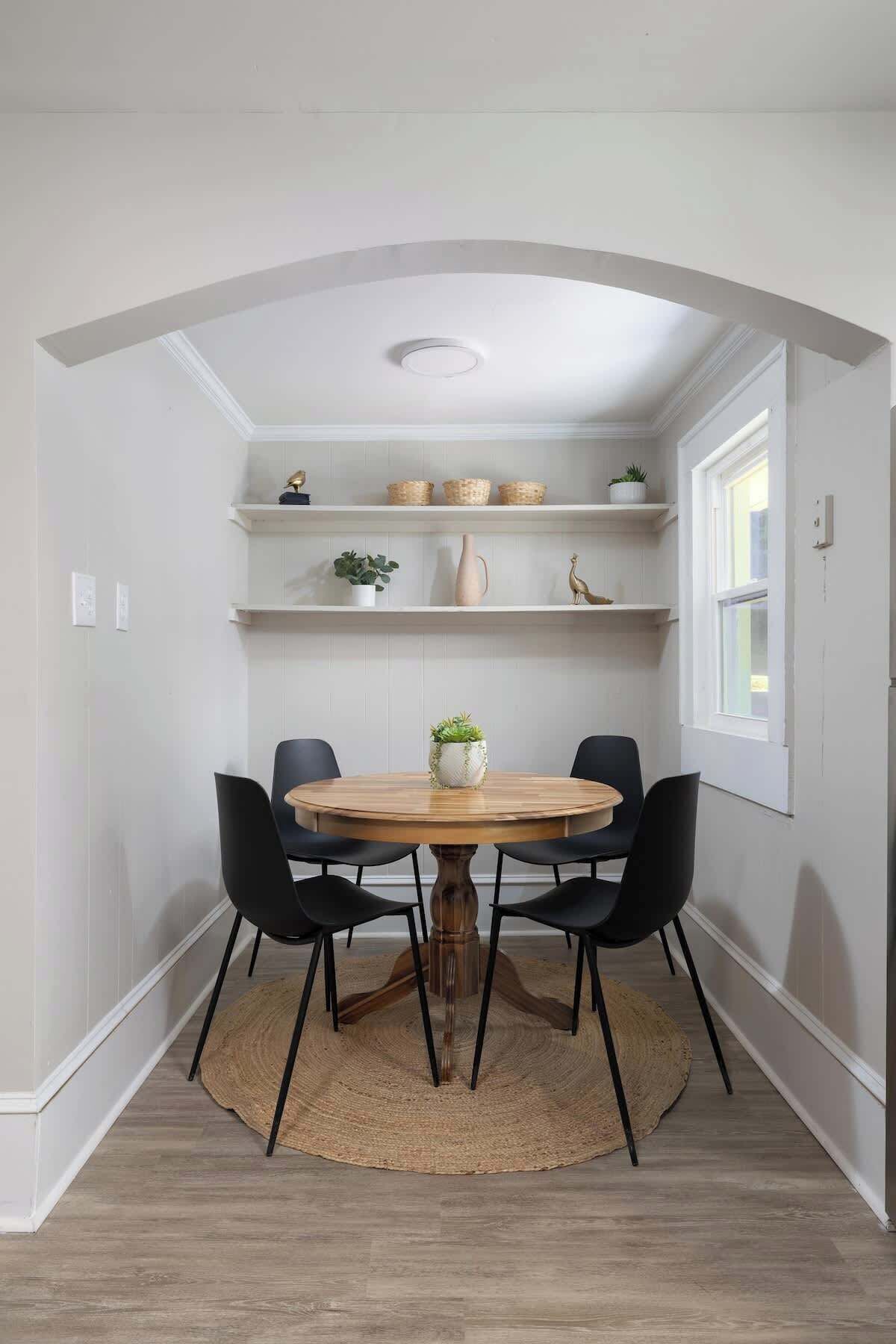 Un coin repas avec une table ronde en bois, quatre chaises noires, un tapis tissé et des étagères ornées de paniers et de plantes.