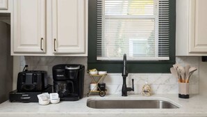 Una cocina con armarios blancos, una cafetera negra, una jarra de café, un fregadero y una ventana con persianas.