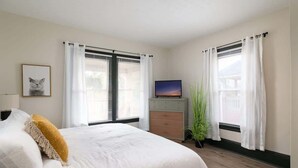 Un dormitorio con cama, televisión, planta y ventana con cortinas.