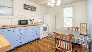Cocina pequeña con microondas, estufa y armarios. Hay una mesa de comedor con mantel a cuadros y una silla de madera.