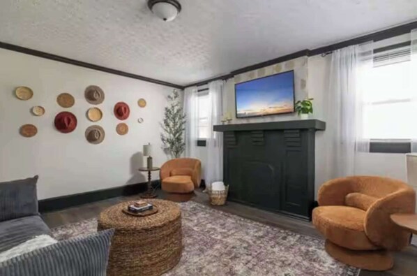 A living room with a fireplace, a sofa, two armchairs, a coffee table, and a wall decorated with hats. - Modern 3BR Executive Suite – Steps from Old Salem & Downtown (Winston-Salem)