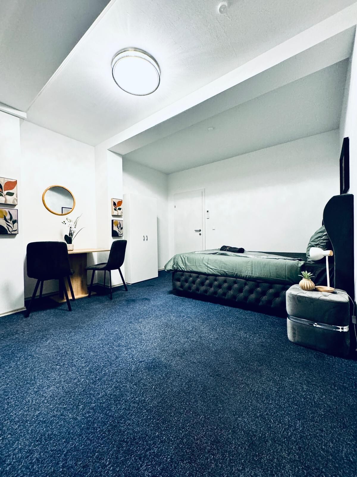 Une pièce compacte et bien éclairée, comprenant un lit, un bureau avec deux chaises, un miroir et une petite table d’appoint avec une lampe.