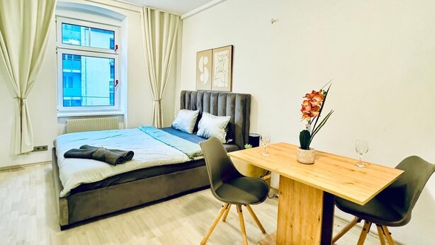 A modern bedroom with a bed, a wooden dining table, two chairs, a window with curtains, and a wall-mounted artwork. - Luxury 7 Bedroom Apartment Sleeps 16 (Vienna)
