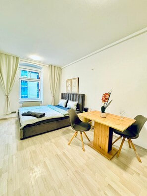 Een moderne slaapkamer met een bed, een houten eettafel, twee stoelen, een raam met gordijnen en een kunstwerk aan de muur.