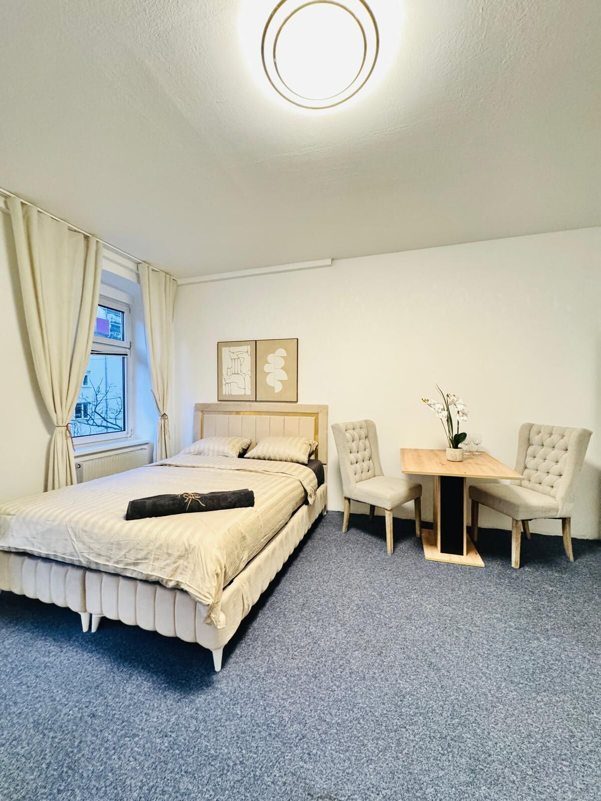A bedroom with a bed, a desk, two chairs, a window with curtains, and a ceiling light.