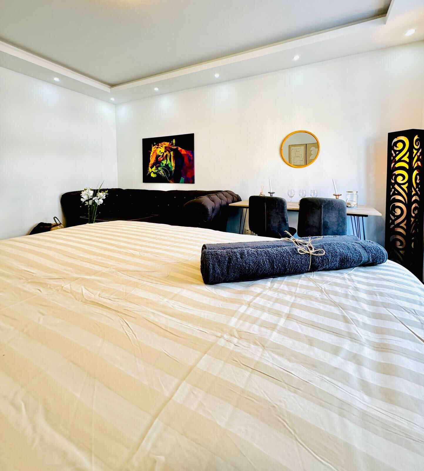 Quarto de hotel moderno com uma cama grande, um sofá preto, um espelho redondo e um objeto decorativo na parede.