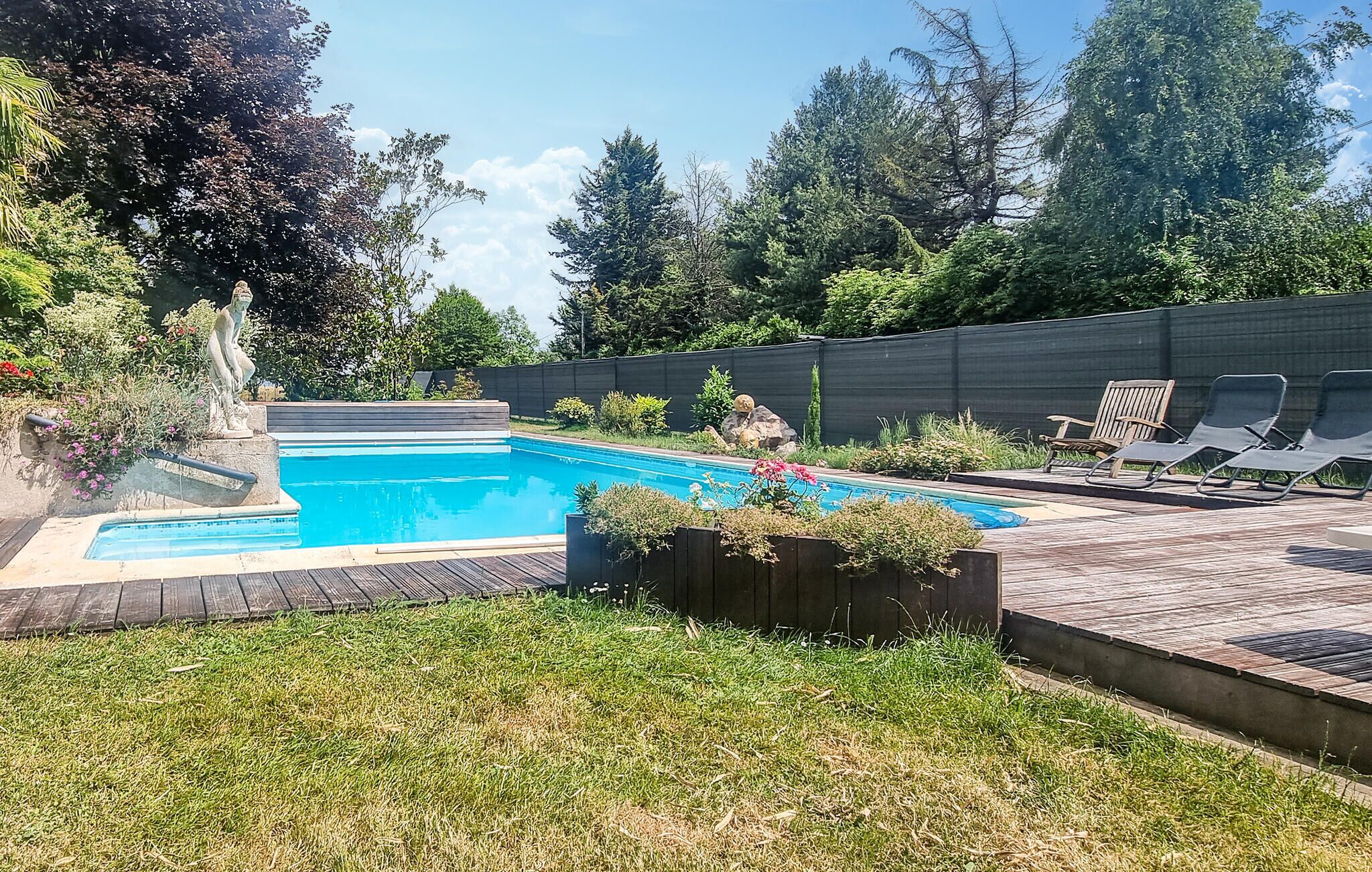 Una piscina rettangolare con una veranda in legno attorno, un'area verde e una recinzione in legno.