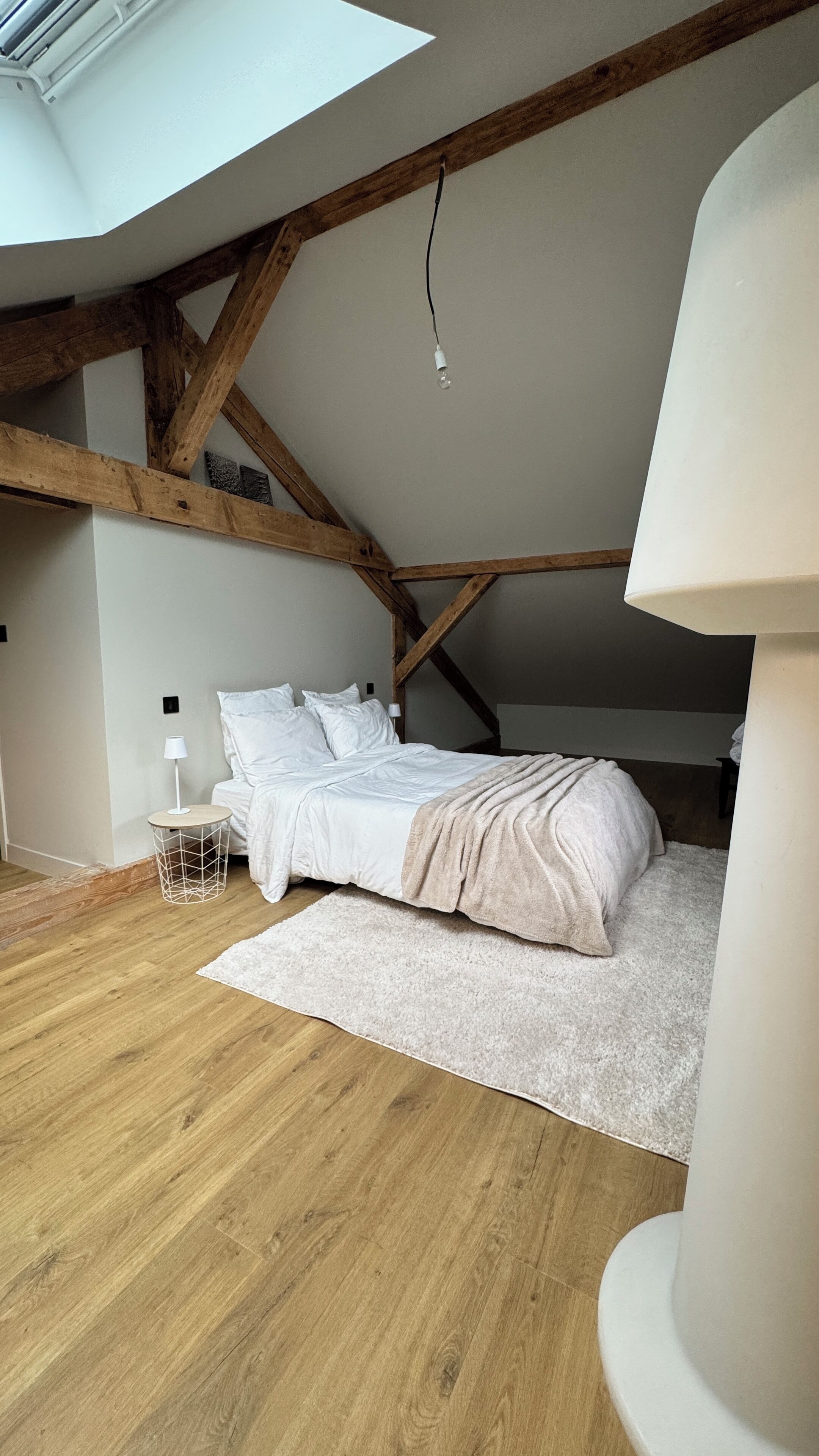 Une chambre avec un plafond en pente, des poutres en bois, un lit avec une literie blanche, une petite table de chevet avec une lampe et un tapis blanc sur le plancher en bois.