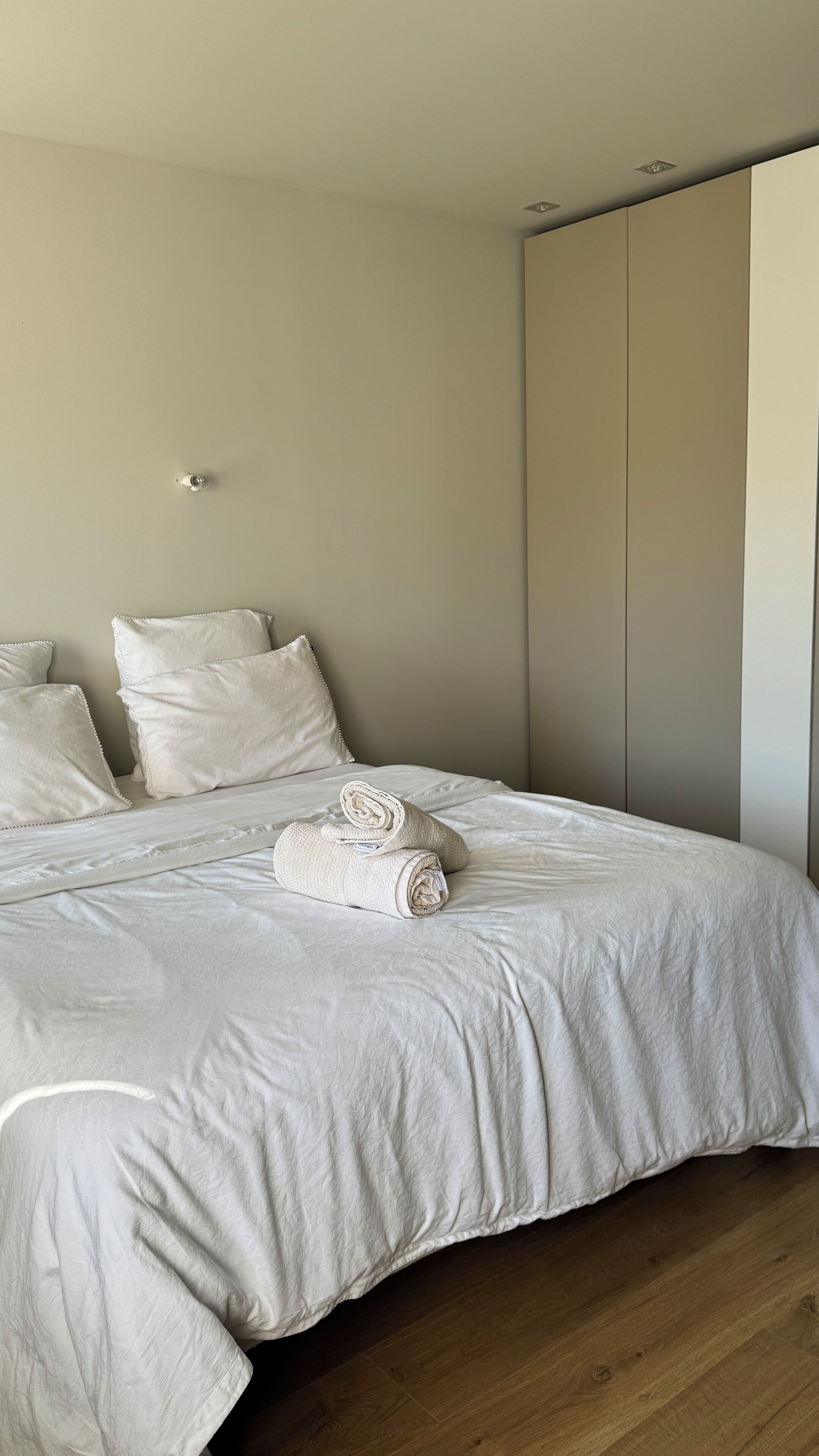 A neatly made bed with white linens and pillows, a wooden floor, and a large wardrobe.