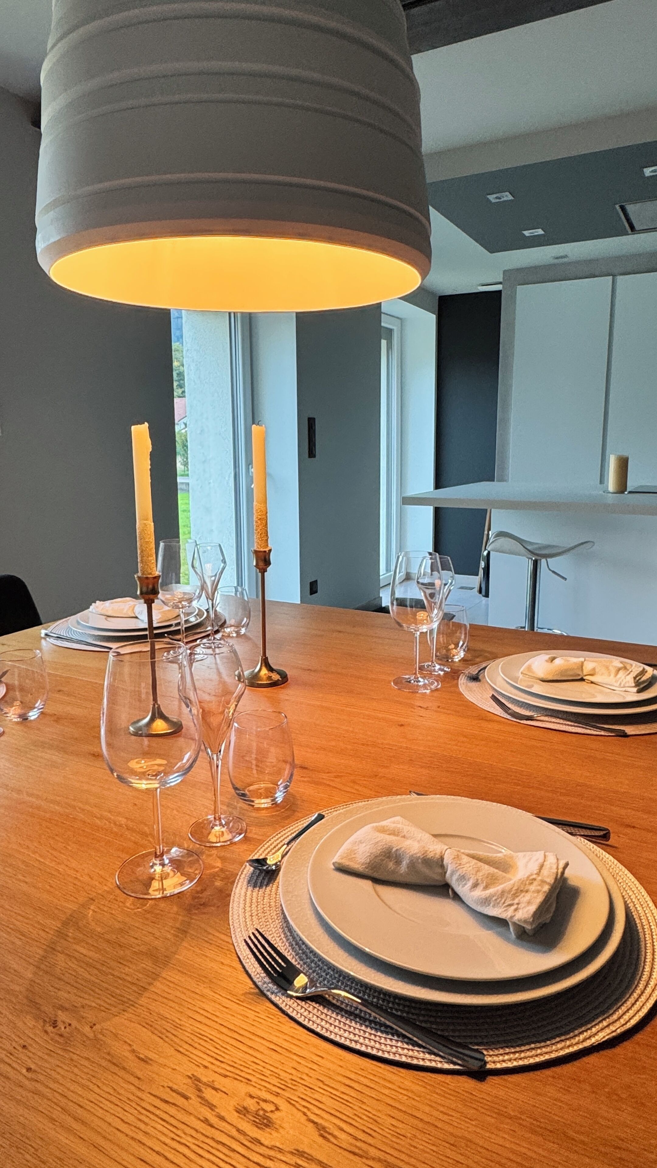 Une table à manger dressée avec des assiettes, des verres et des couverts, agrémentée de bougies et d’un luminaire suspendu au design moderne.