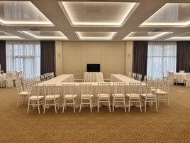 A conference room with a long table surrounded by chairs, a television, and large windows with curtains.