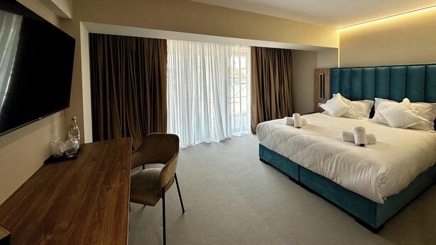 A hotel room with a bed, a desk, a chair, a TV, and curtains. - River Park Dragasani (Drăgășani)
