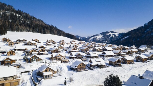 Ein verschneites Dorf mit Holzhütten, blauem Himmel und Bergen im Hintergrund.