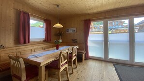 Ein Holzblockhaus-Interieur mit Esstisch, Stühlen, einer Bank, einer Pendelleuchte und großen Fenstern mit Vorhängen.
