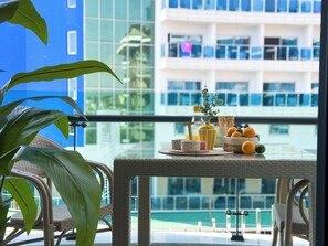 Ein Balkon mit Korbmöbeln, einem Tisch mit Früchten und einer Vase, sowie ein modernes Gebäude im Hintergrund.