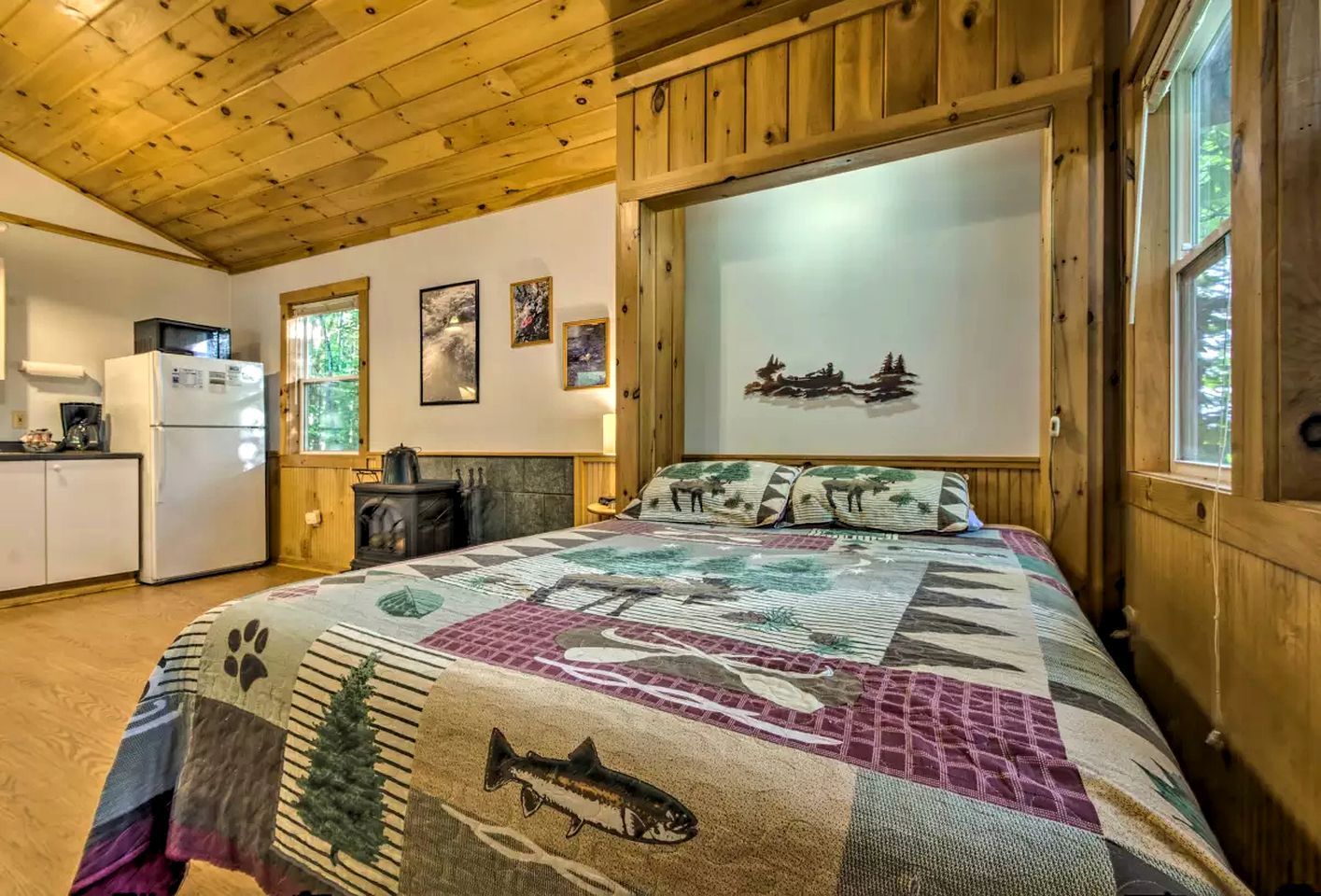 A cozy cabin interior with a wooden ceiling, a bed with a quilt, a fireplace, a kitchen area, and a wall with framed pictures.
