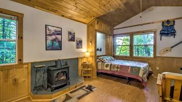 A cozy cabin room with a wood stove, a bed with a patterned blanket, and a view of the forest through large windows.