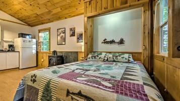 Een gezellig houten interieur met een bed met patchworkdeken, een open haard, een keukenhoek en een wand met ingekaderde foto's.
