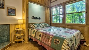 A wooden cabin bedroom with a bed, a nightstand, a lamp, and a window with a forest view. - Tree-House Style Cabin with Forest Views, Nantahala National Fores, North Carolina (Bryson City)