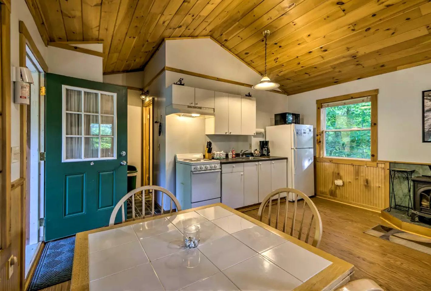 A compact kitchen with a stove, refrigerator, and cabinets, a dining table with chairs, and a window with a view of greenery.
