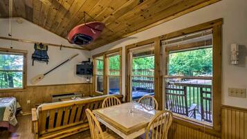 A cabin interior with wooden walls, a dining table, chairs, a bench, a ceiling fan, and large windows with a view of the outdoors.