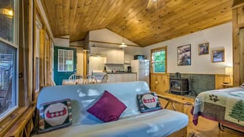 A cozy cabin interior with a wooden ceiling, a kitchenette, a sofa with decorative pillows, and a fireplace.