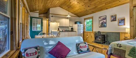 A cozy cabin interior with a wooden ceiling, a kitchenette, a sofa with decorative pillows, and a fireplace.
