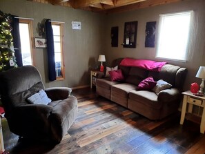 A cozy living room with a brown leather armchair, a brown sofa with a pink blanket, a wooden floor, and a window with blinds.