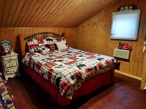 A bedroom with a wooden ceiling, a bed with a checkered quilt, a nightstand with a decorative item, and a window with blinds.