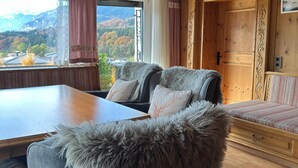 A cozy room with a wooden table, two cushioned chairs with fur trim, a sofa with a patterned cushion, and a view of mountains through a window. - Cozy holiday home with mountain views (Breitenbach am Inn)