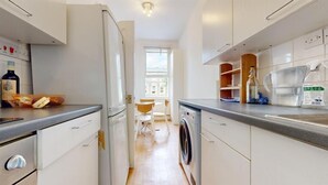 A modern kitchen with white cabinets, a washing machine, and a dining area with a table and chairs. - Philbeach Gardens Flat (London)
