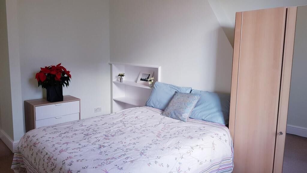 A bedroom with a bed, a bedside table with a vase of flowers, a wardrobe, and a shelf with decorative items.