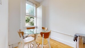 A dining area with a round glass table and six chairs, a window with a view of trees, and a wooden floor. - Philbeach Gardens Flat (London)