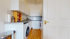 A compact kitchen with white cabinets, a washing machine, and a countertop with various items. - Philbeach Gardens Flat (London)