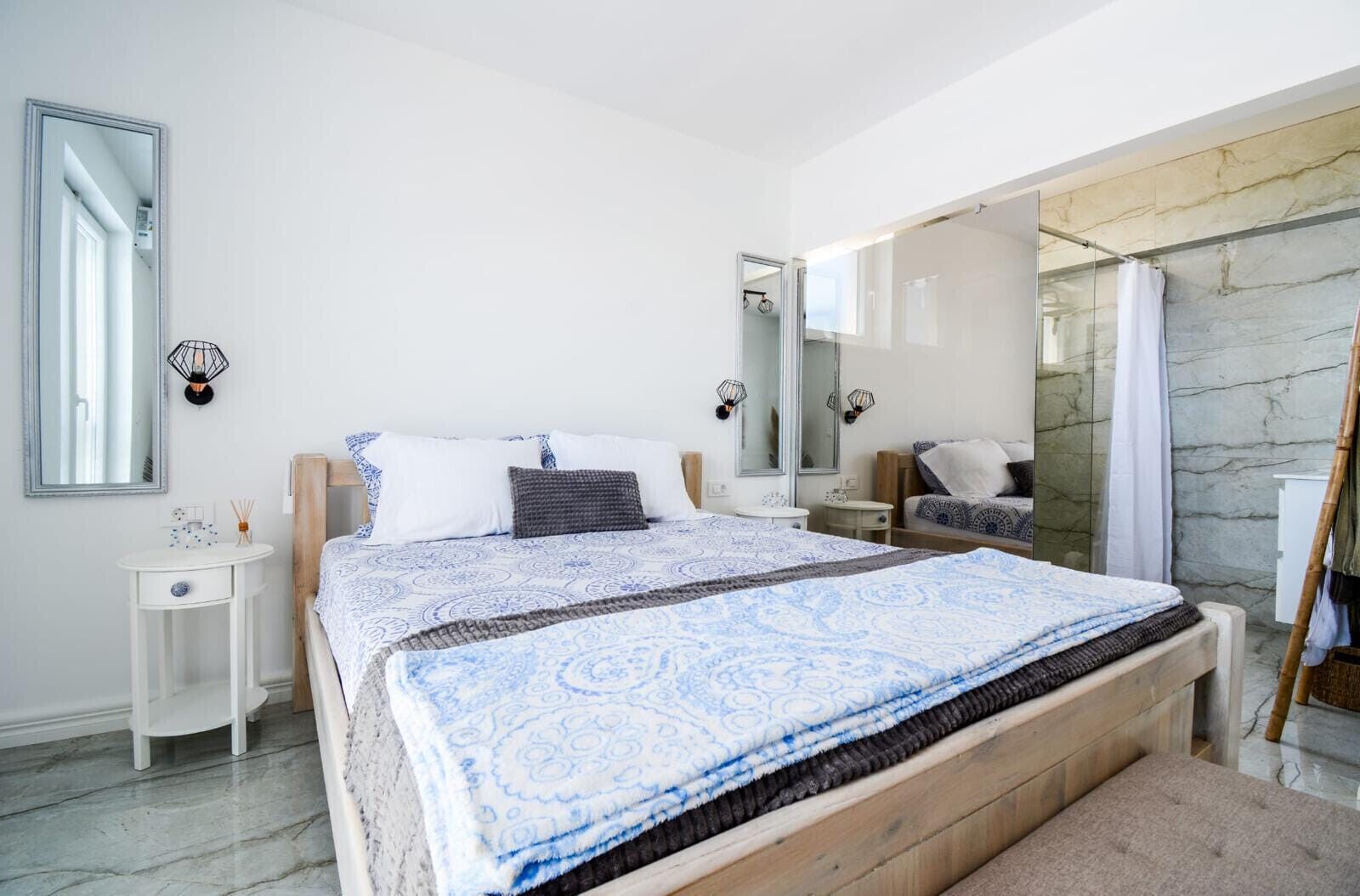 Un dormitorio moderno con una cama grande, mesitas de noche, un espejo y un baño con ducha y paredes de mármol.
