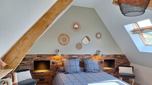 Een slaapkamer met een schuine kap, een metselwerk open haard, twee nachtkastjes met lampen, een bed met een blauw geruit dekbed en een dakraam.