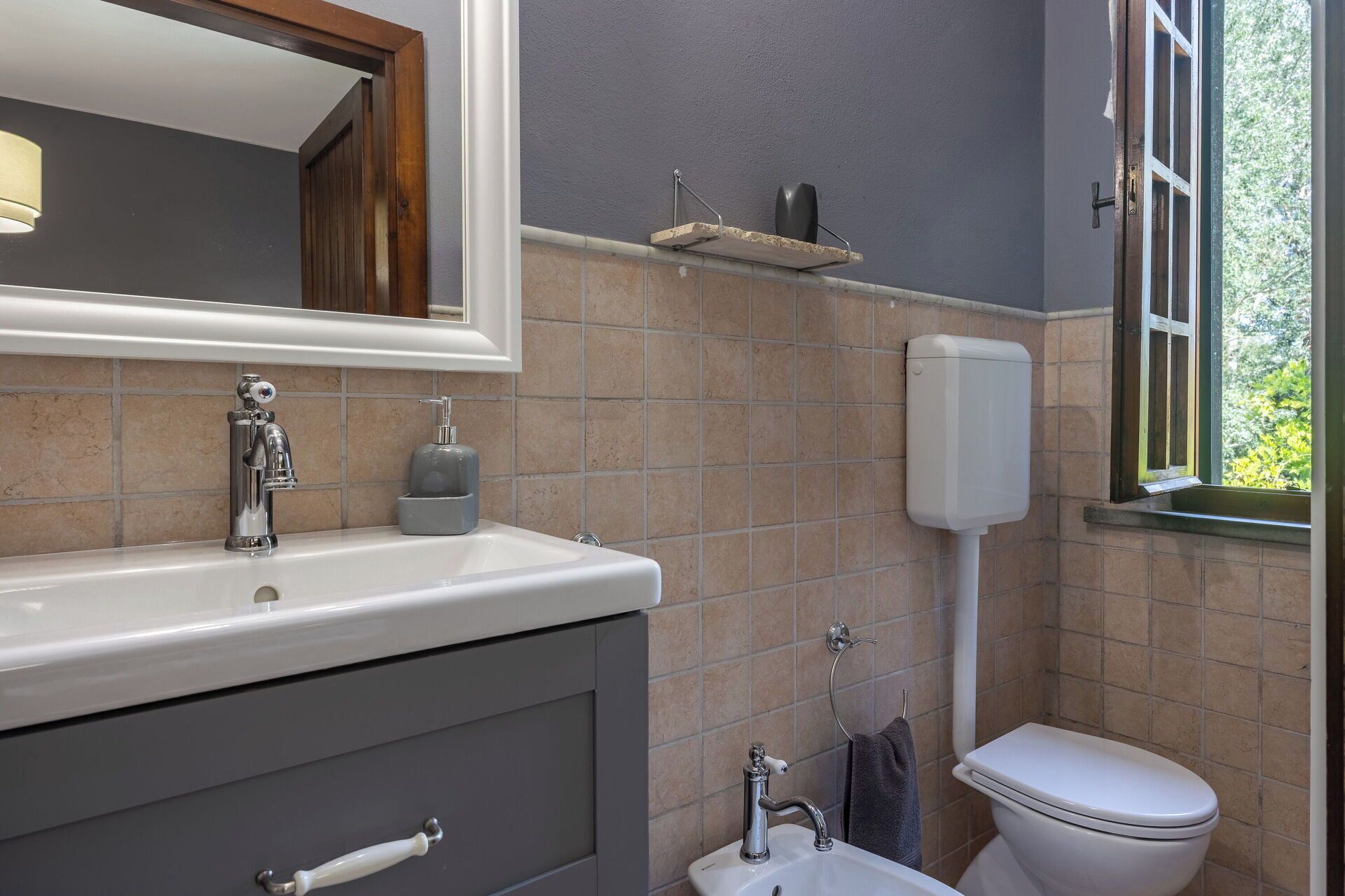 Une salle de bain avec un lavabo blanc, un miroir, des toilettes et une fenêtre donnant sur un espace verdoyant.