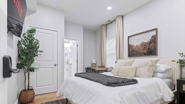 Un dormitorio con cama, mesita de noche, lámpara, planta, puerta y televisor fijado a la pared.