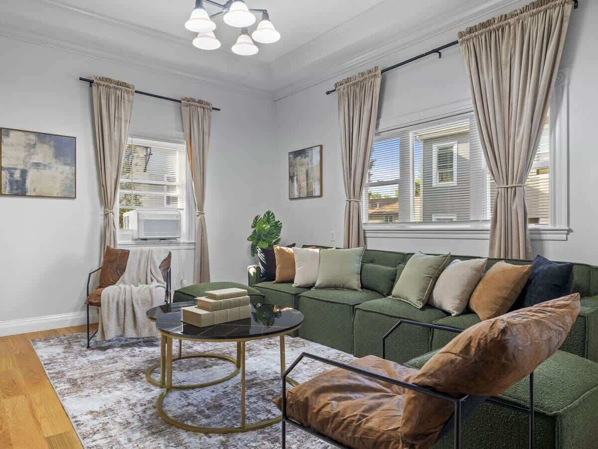 Een woonkamer met een groene bank, een ronde salontafel en een stoel. Er hangen gordijnen voor de ramen, en op de vensterbank staat een plant.