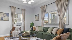 Una sala de estar con un sofá verde, una mesita de centro redonda y una silla. Hay cortinas en las ventanas, y una planta en el alféizar.