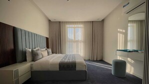 A modern hotel room with a large bed, a bedside table, a small ottoman, a built-in wardrobe, and a large window with curtains. - Velmist Residence Abha (Abha)
