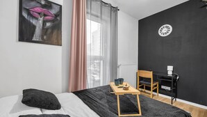 Un dormitorio con cama, mesita de noche con bandeja, una silla, escritorio y un reloj de pared.