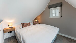 Ein Schlafzimmer mit schrägverlaufender Decke, einem Bett mit weißer Bettwäsche, gemusterten Kissen, einem Nachttisch mit Lampe und einem Fenster mit Rollo.