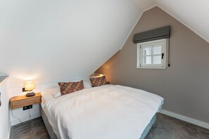 Ett sovrum med ett sluttande tak, en säng med vita sängkläder, mönstrade kuddar, ett sängbord med en lampa och ett fönster med en fönsterlucka.