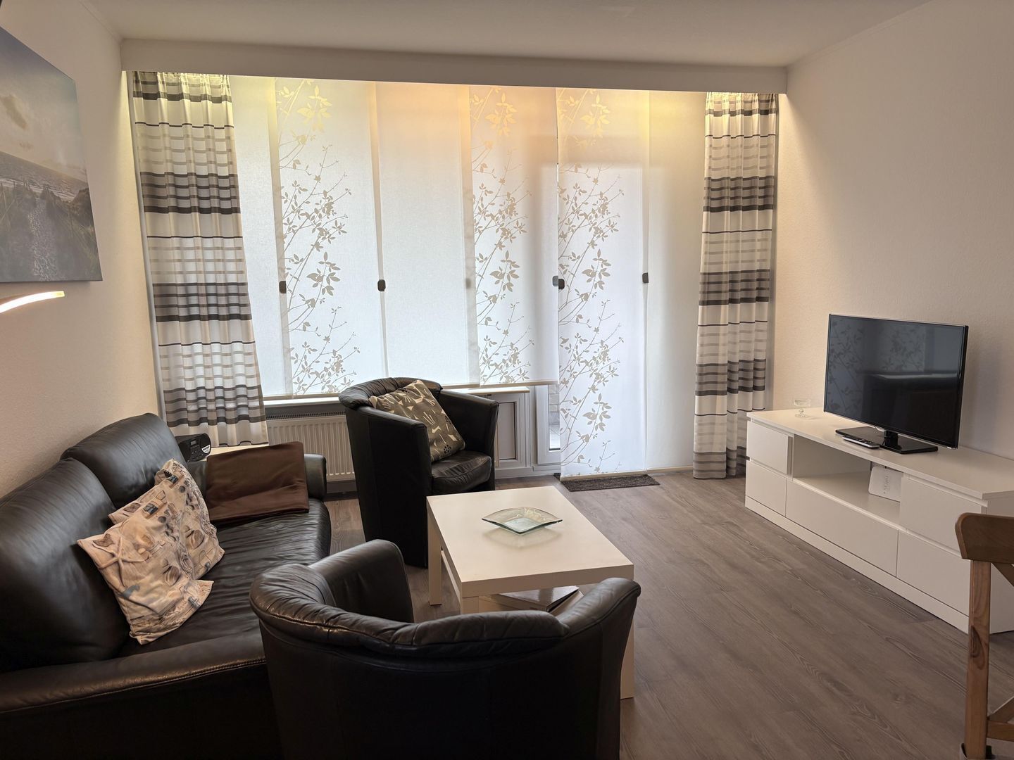 A modern living room with a sofa, armchair, and coffee table. There is a TV on a stand and a window with curtains.