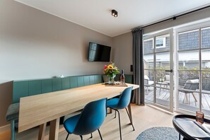 A modern dining area with a wooden table, blue chairs, a bench, and a TV mounted on the wall. - Dependance Windrose 172 - Dependances Windrose (Wenningstedt-Braderup (Sylt))