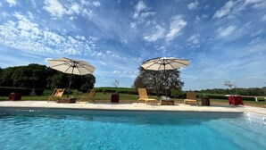 Une piscine entourée d’un espace aménagé avec des arbres, des chaises longues et des parasols, le tout sous un ciel dégagé.