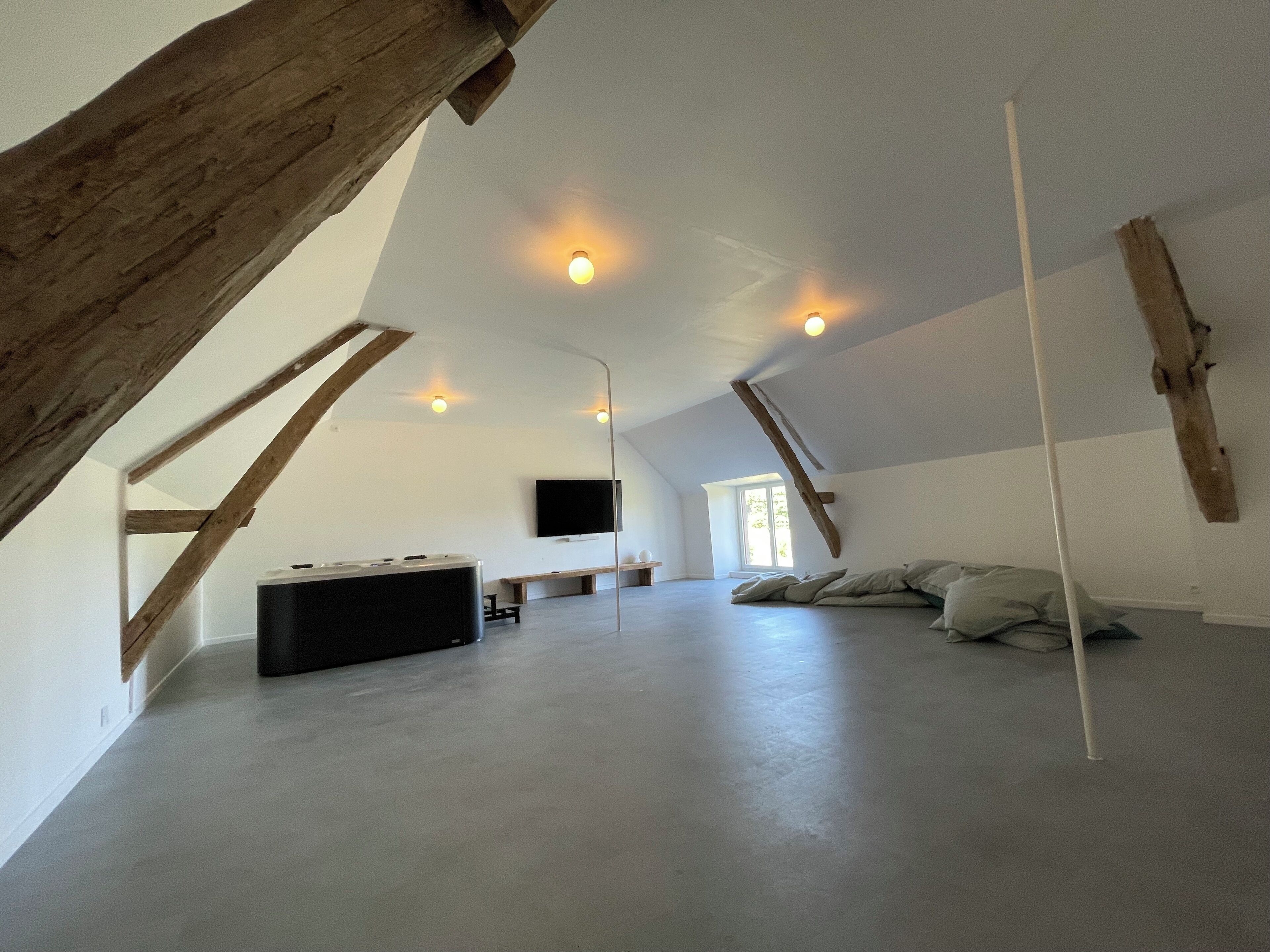 Amplia habitación en ático con techo alto, vigas de madera a la vista, un ventanal grande, televisor de pantalla plana y un sofá.