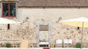 Un bâtiment en pierre avec un toit en tuiles, des tables à l’extérieur sous des parasols et un espace piscine.